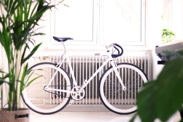 white bicycle in front of a large radiator with two houseplants in the foreground.